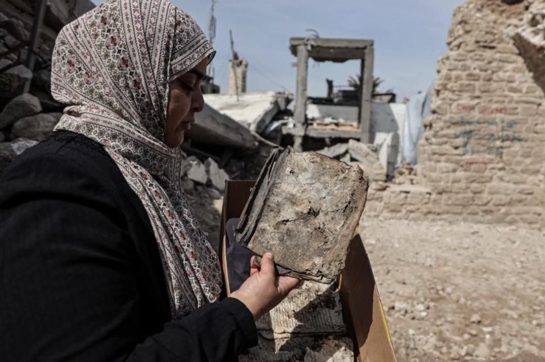 Una donna con un foulard sul capo tiene tra le mani un libro gravemente danneggiato e ricoperto di polvere. Sullo sfondo sono visibili macerie e resti di edifici distrutti.