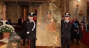 Due Carabinieri in alta uniforme trasportano un dipinto a soggetto religioso all’interno di una chiesa. L’opera raffigura la Madonna con il Bambino ed è circondata da decorazioni e composizioni floreali.