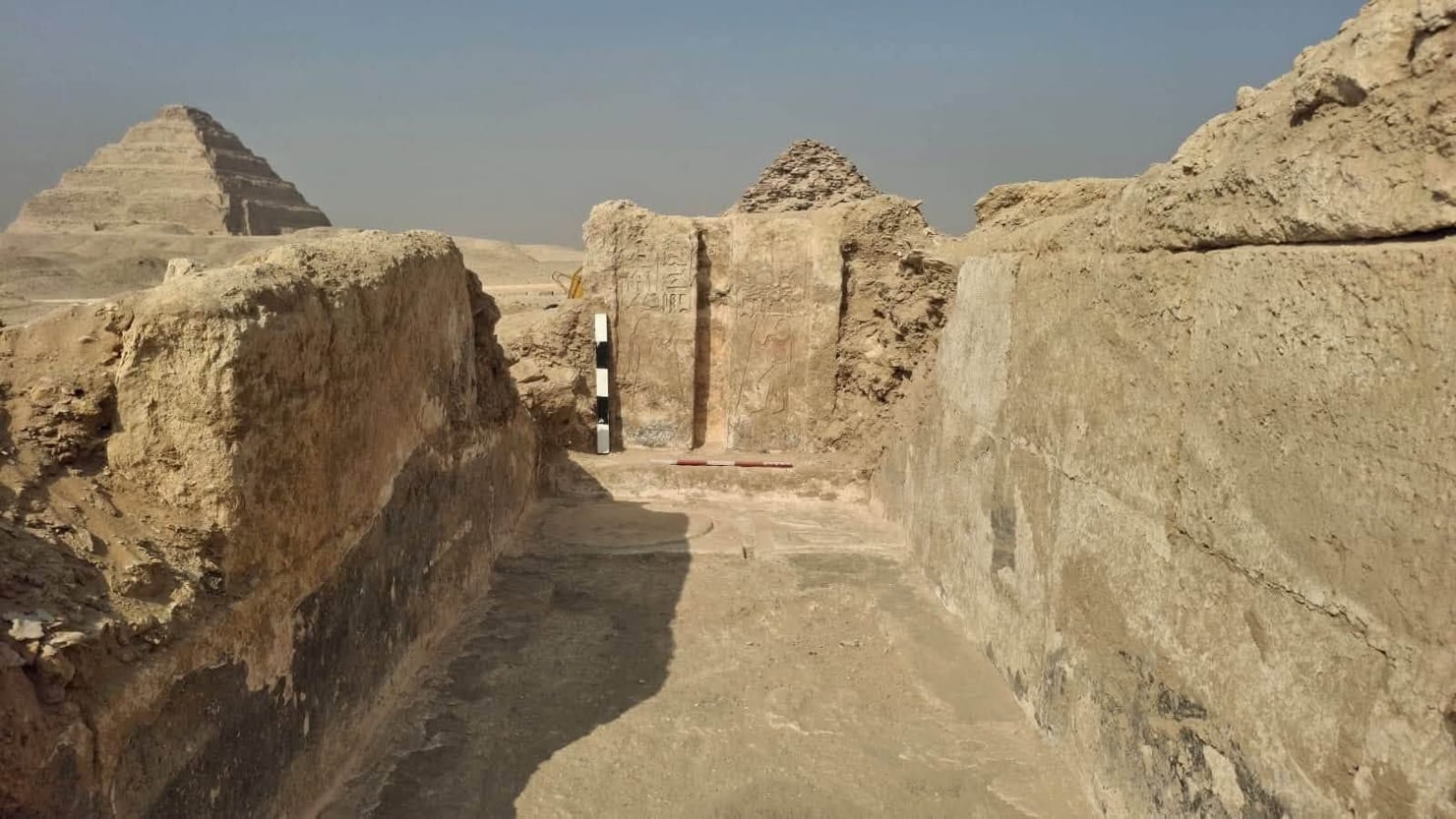 Area archeologica di Saqqara con i resti di antiche strutture in pietra e mattoni. Al centro si vede una porta scolpita nella pietra, priva di un passaggio oltre di essa; sullo sfondo emerge una piramide nella distesa desertica.