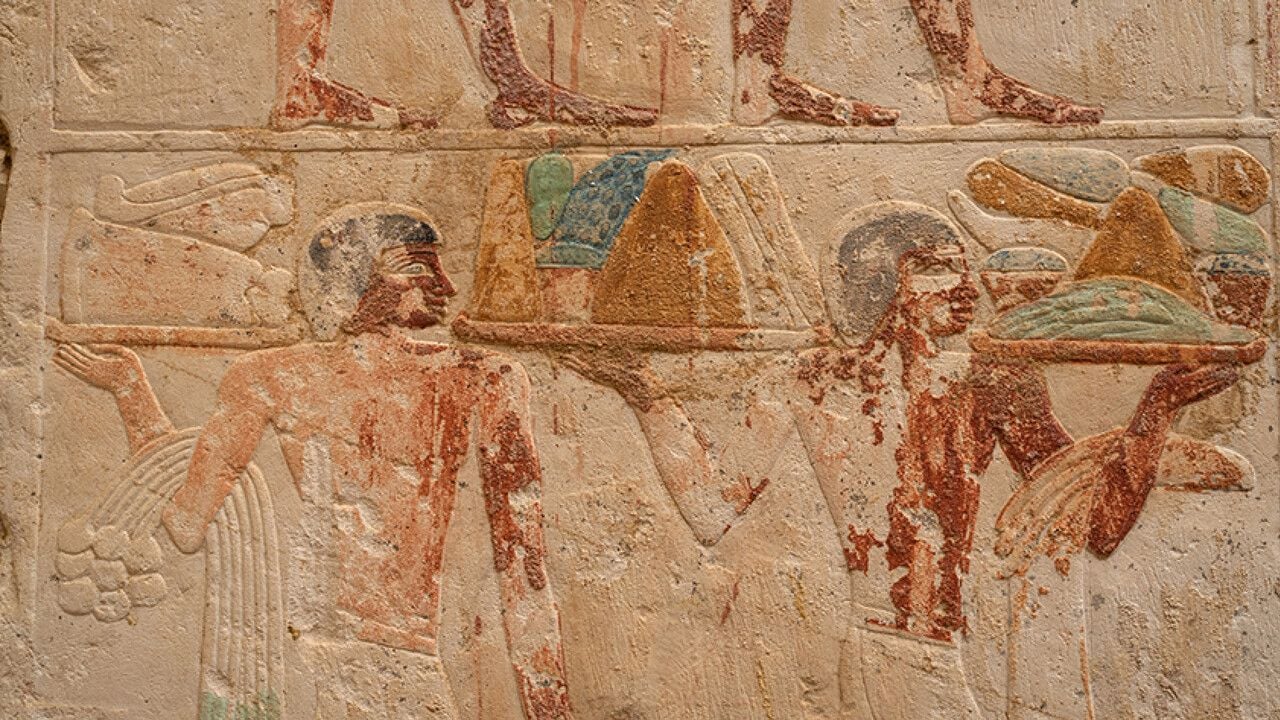 Rilievo dell’antico Egitto raffigurante due uomini di profilo, vestiti con tuniche e copricapi, mentre trasportano un grande recipiente decorato. La scena è scolpita nella pietra e conserva tracce di colore rosso, blu e verde.