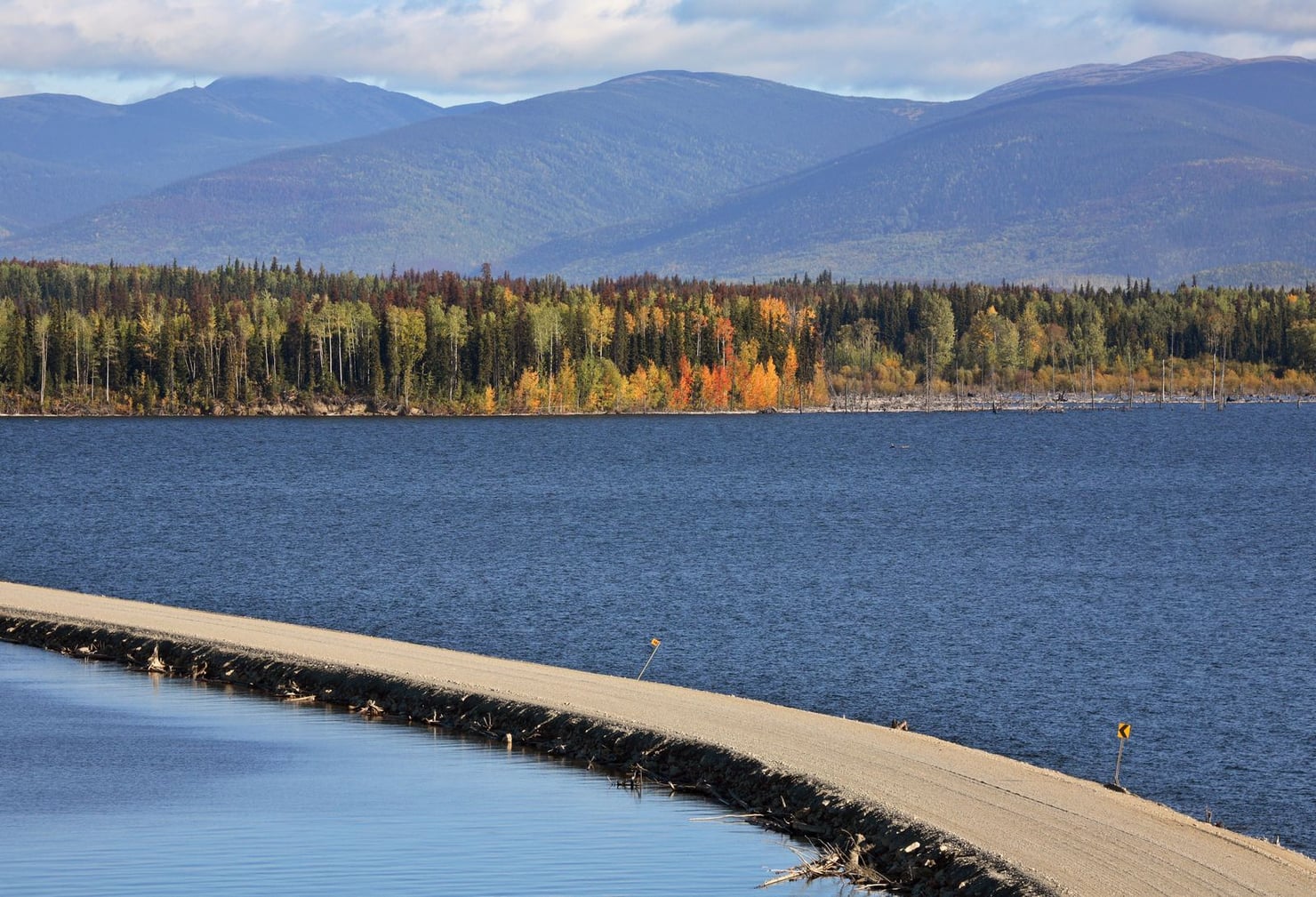 Paesaggio autunnale con un lago in primo piano, una strada rialzata che attraversa l’acqua e una fascia di bosco dai colori gialli, arancioni e verdi. Sullo sfondo si estende una catena montuosa sotto un cielo nuvoloso.