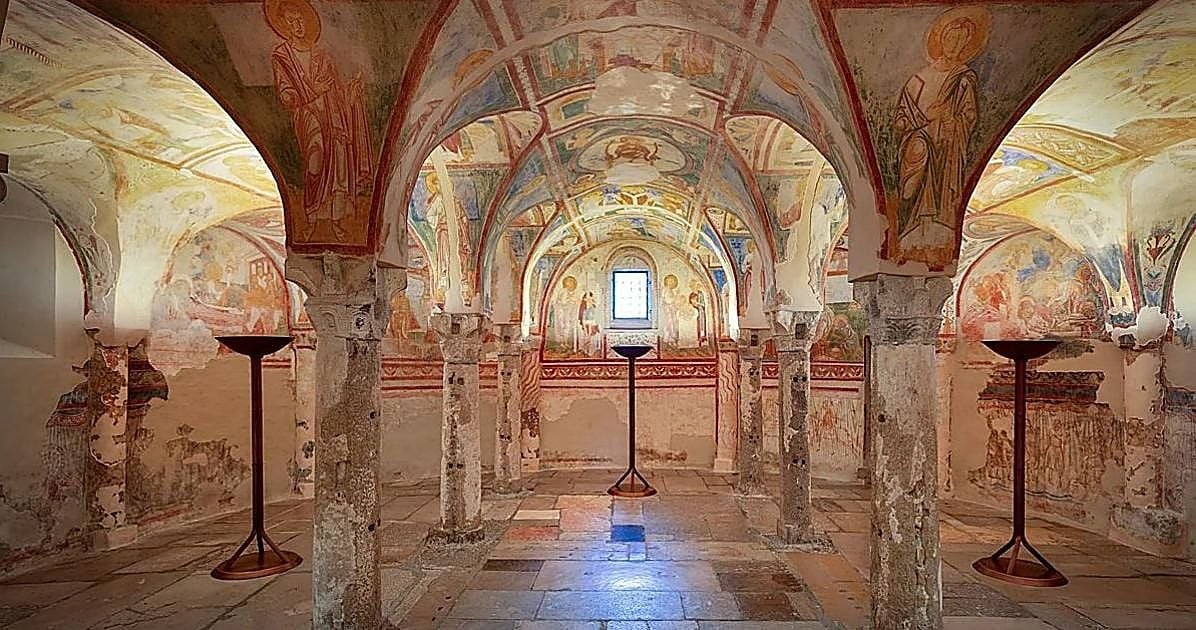 Interno di una cappella medievale con volte e colonne in pietra, interamente decorata da affreschi con figure religiose e motivi ornamentali. Al centro della sala si trova un piccolo altare, mentre il pavimento è composto da lastre di pietra di diversi colori.