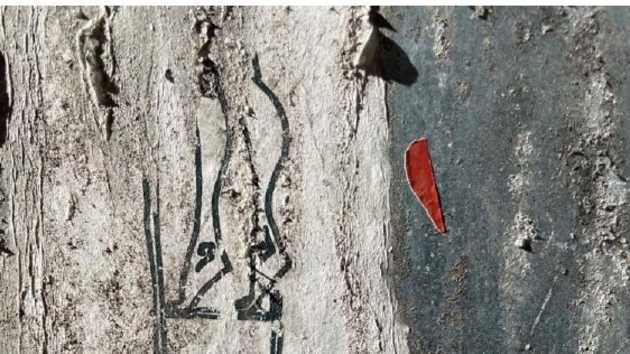 Dettaglio di una superficie muraria chiara e irregolare, con un disegno realizzato in linee nere che raffigura una figura umana stilizzata. A destra del disegno è visibile un piccolo elemento rosso, simile a una foglia o a un frammento di carta.
