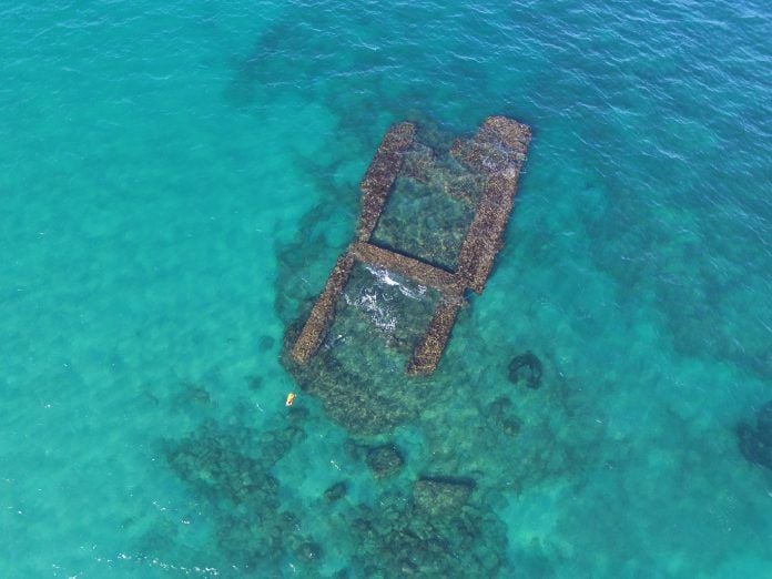 Veduta dall’alto di una struttura archeologica sommersa nelle acque turchesi del mare. I resti, ricoperti di vegetazione marina, sono visibili attraverso l’acqua limpida e presentano una forma rettangolare con due strutture laterali.