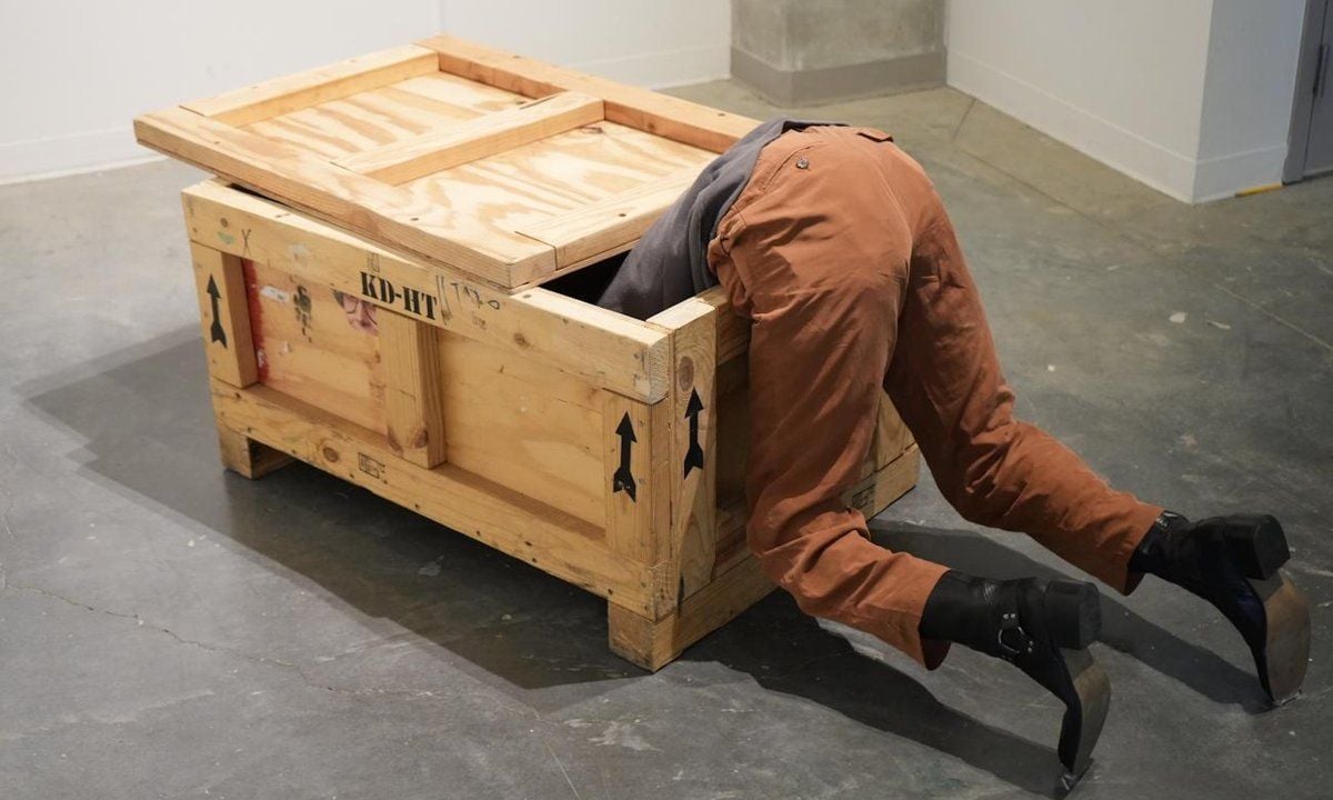 Installazione artistica che raffigura una persona con il busto immerso all’interno di una grande cassa di legno, mentre le gambe, vestite con pantaloni marroni e stivali neri, sporgono all’esterno. L’opera è esposta in uno spazio museale dal pavimento in cemento.