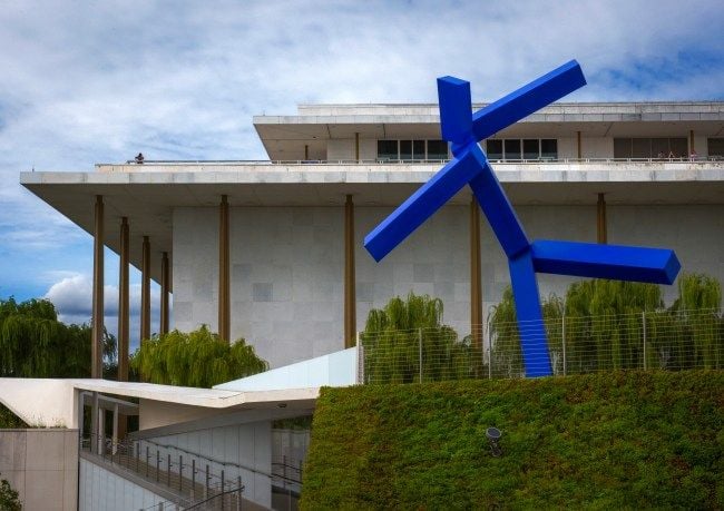 A bright blue sculpture resembling a dancing stick figure looms above a modern white building, with green bushes in the foreground.