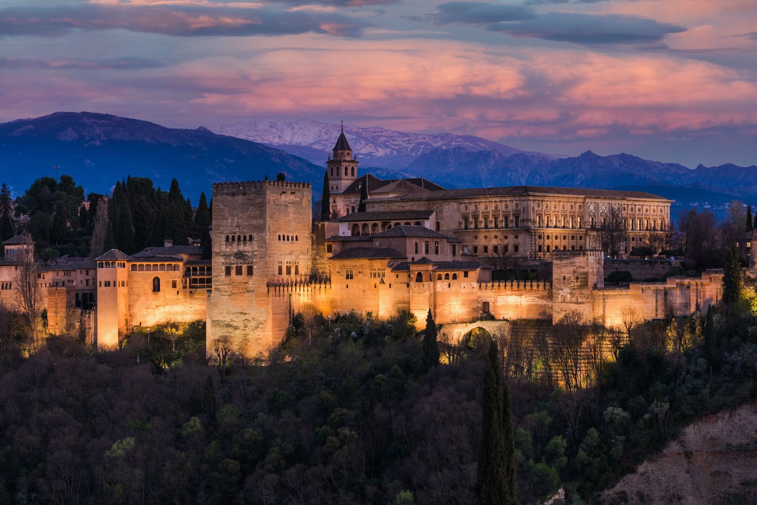 Veduta panoramica dell’Alhambra di Granada al tramonto, con le mura e gli edifici del complesso illuminati dalla luce calda della sera. Sullo sfondo si estende la Sierra Nevada, con le montagne parzialmente innevate sotto un cielo nuvoloso.
