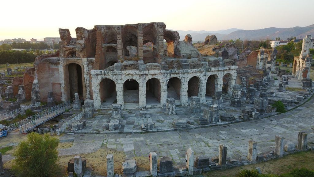 Veduta di un sito archeologico con i resti di un antico edificio in pietra e mattoni, caratterizzato da una fila di archi. Intorno sono visibili colonne e altri elementi architettonici in rovina, con montagne sullo sfondo.