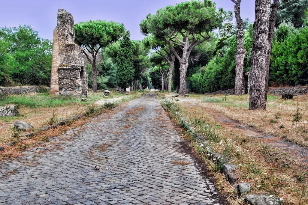 Antica strada romana lastricata in pietra, fiancheggiata da pini e vegetazione. Ai lati del percorso sono visibili resti di strutture e monumenti in muratura.