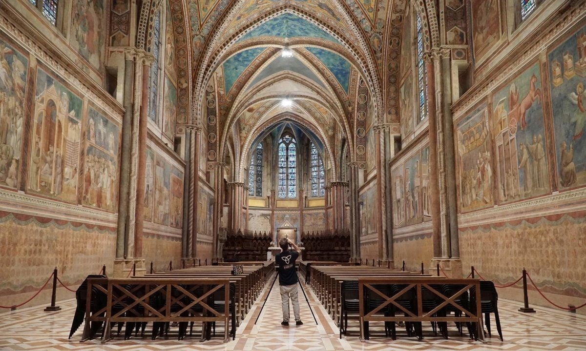Interno di una basilica medievale con alte volte a crociera e pareti interamente decorate da cicli di affreschi. File di sedute occupano la navata centrale, mentre una persona in piedi al centro fotografa l’interno. Sul fondo sono visibili l’altare e alte vetrate.