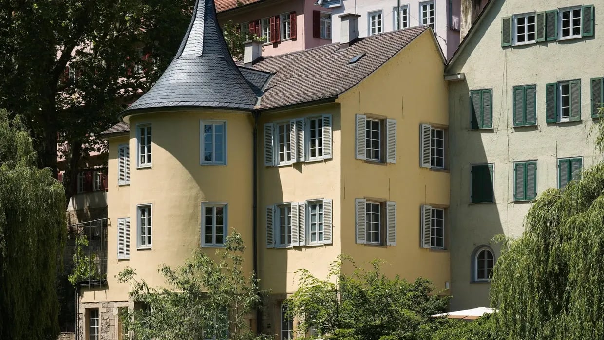 Veduta esterna di un edificio storico dalle pareti gialle, con una torretta circolare dal tetto conico e numerose finestre con persiane chiare. La costruzione è circondata da alberi e vegetazione e affiancata da un altro edificio con persiane verdi.