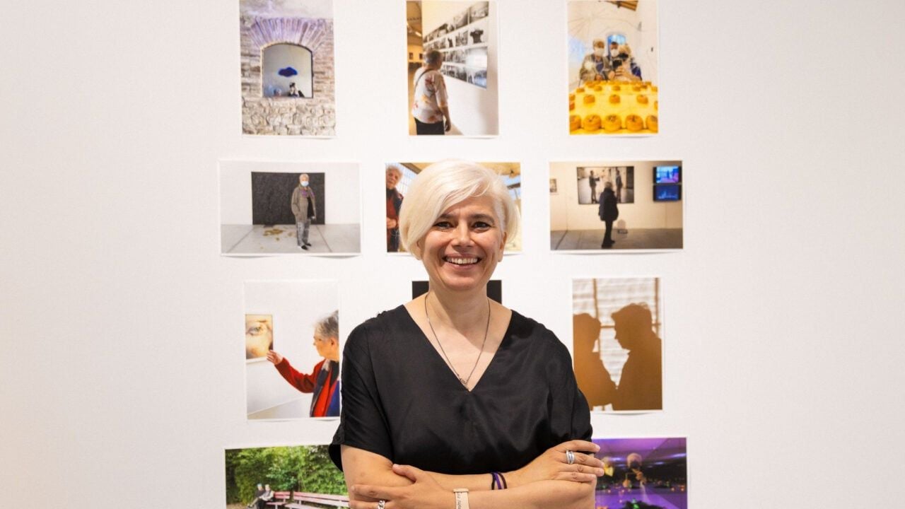 Ritratto fotografico di una donna con capelli corti e bianchi, sorridente e con le braccia incrociate, davanti a una parete sulla quale sono esposte diverse fotografie e immagini di una mostra.