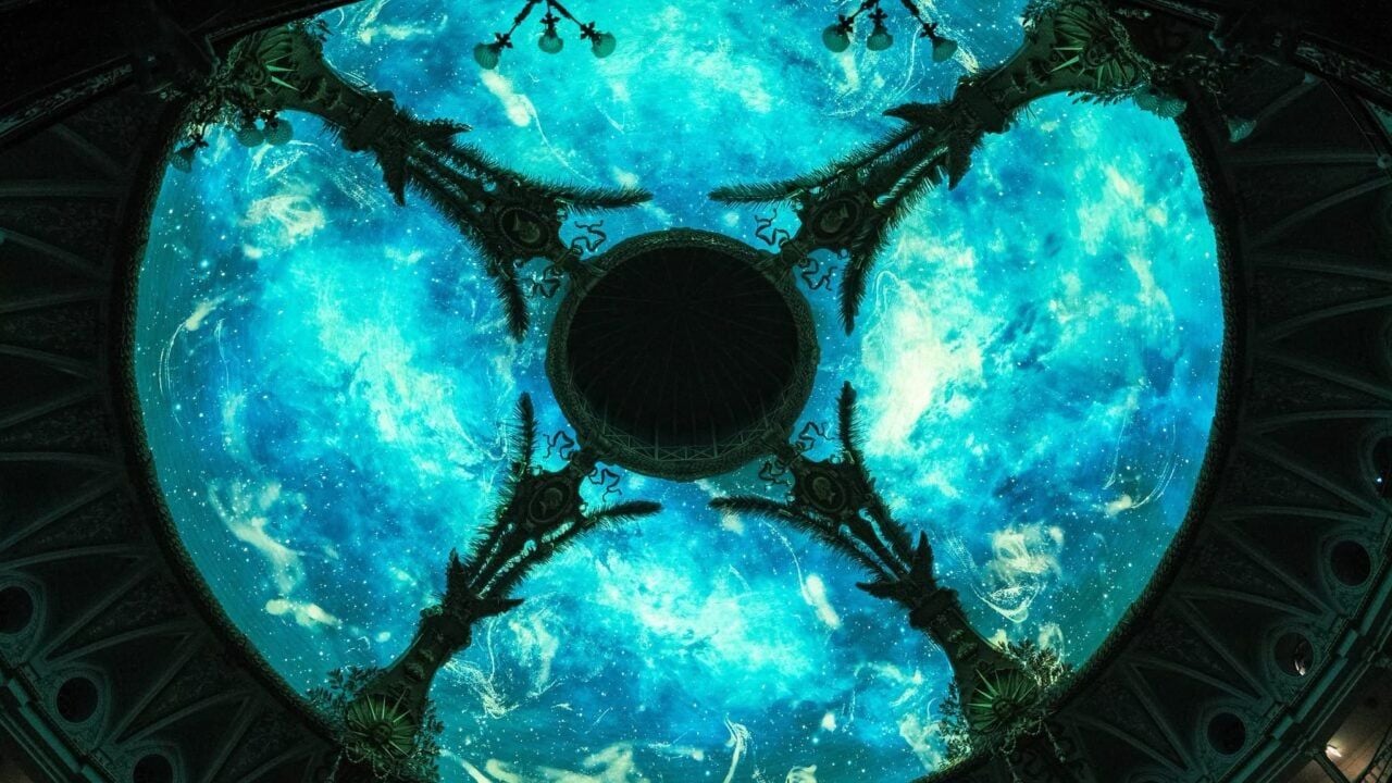 Veduta dal basso di una grande cupola suddivisa in quattro sezioni, illuminate da una proiezione digitale nei toni dell’azzurro e del turchese. Le immagini evocano un cielo o uno spazio cosmico, incorniciato dagli elementi architettonici e decorativi della cupola.