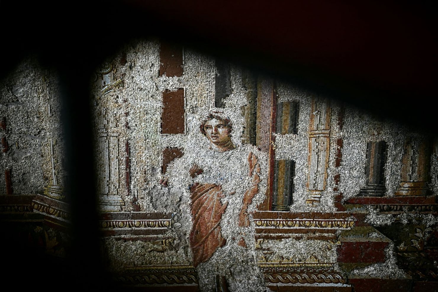 Un dettaglio di un mosaico romano raffigura una figura umana seduta, circondata da edifici e decorazioni architettoniche. La superficie del mosaico presenta numerose lacune e parti deteriorate.