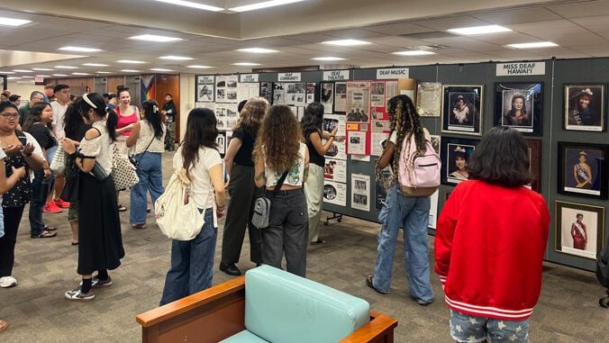 Persone visitano una mostra allestita in una sala, osservando pannelli informativi, fotografie e opere dedicate alla cultura e alla comunità sorda. In primo piano, una persona osserva l’esposizione; sullo sfondo sono visibili pannelli con le scritte “Deaf Community”, “Deaf Music” e “Miss Deaf Hawaii