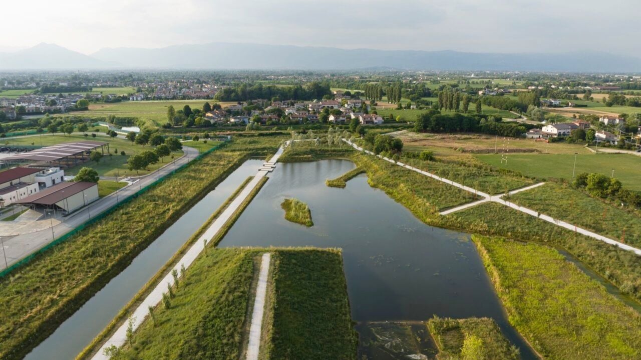 Vista aerea del Parco della Pace di Vicenza, con un ampio specchio d’acqua attraversato da percorsi pedonali, aree verdi e vegetazione. Sullo sfondo si estende il paesaggio agricolo e urbano, con le montagne all’orizzonte.
