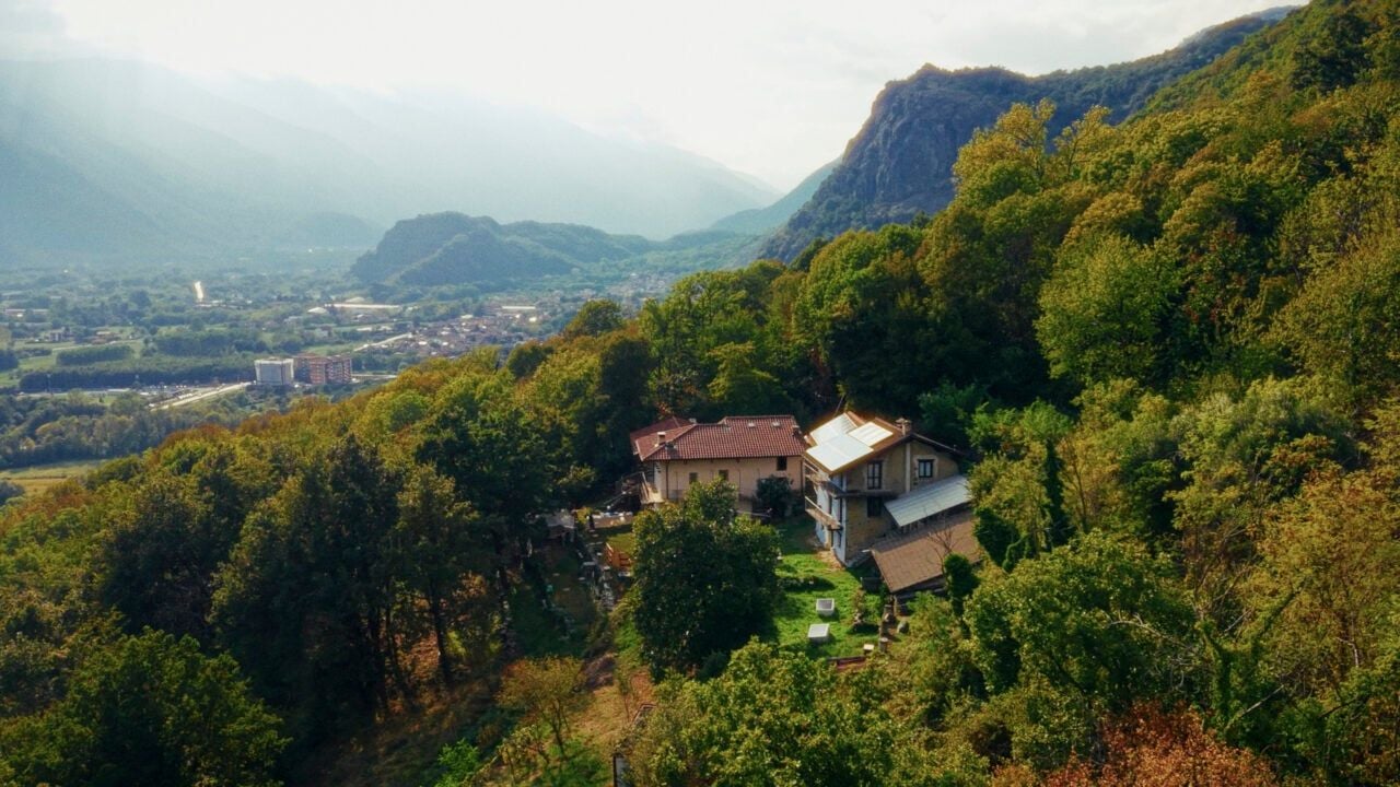 Veduta dall’alto di alcuni edifici immersi in un fitto bosco su un versante montano. Sullo sfondo si apre una valle con un centro abitato, circondata da rilievi montuosi.
