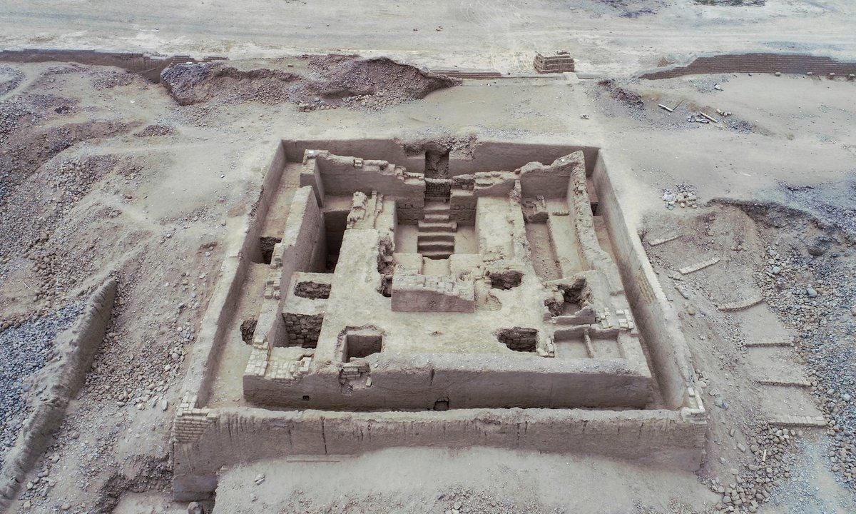 Veduta aerea di uno scavo archeologico in un’area desertica. Al centro sono visibili i resti di una struttura rettangolare, con muri in mattoni, diversi ambienti interni, aperture e una scala che conduce a un livello inferiore. Intorno emergono altre tracce di strutture e murature.