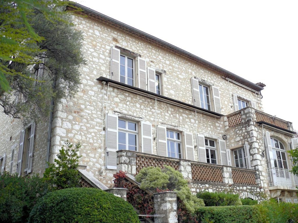 Veduta esterna di una villa in pietra chiara, con due piani, persiane bianche e ampie finestre. Una terrazza con balaustre in pietra si affaccia sul giardino, ricco di siepi e piante ornamentali.