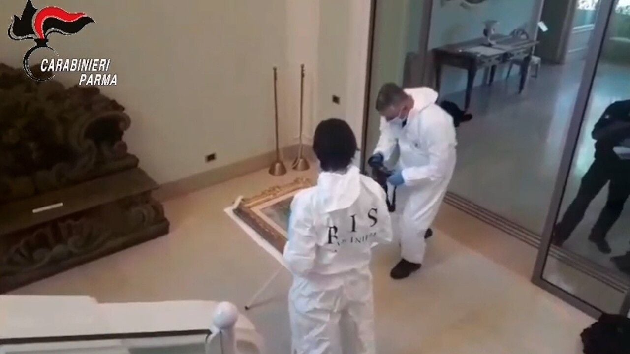 Due operatori in tute bianche e guanti esaminano un dipinto appoggiato a terra all’interno di un edificio. Sullo sfondo si vedono alcune sale e arredi. In alto a sinistra è presente il logo dei Carabinieri Parma.