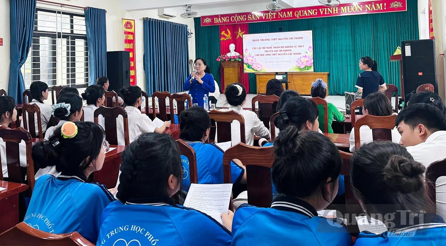 Incontro in un’aula scolastica in Vietnam, con numerosi studenti seduti ai banchi. Davanti alla classe, una donna parla al microfono, mentre alle sue spalle sono visibili un palco, uno schermo e simboli istituzionali vietnamiti.