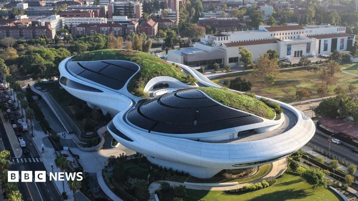 Veduta aerea del Museo Lucas dalle forme sinuose, con ampie coperture scure e tetti ricoperti di vegetazione. L’edificio è circondato da aree verdi e si trova in un contesto urbano, con altri edifici e strade visibili sullo sfondo.