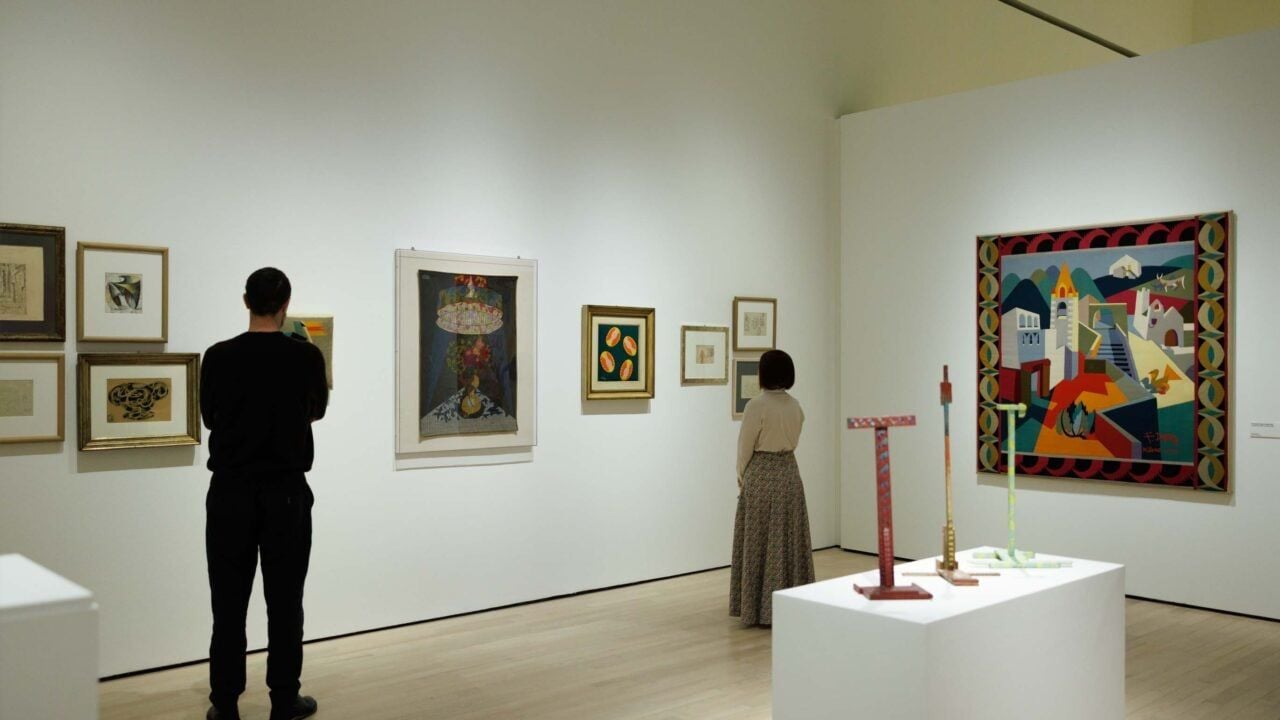 Sala espositiva di un museo con dipinti, opere grafiche e sculture contemporanee disposti sulle pareti e su un piedistallo centrale. Due visitatori osservano le opere esposte.