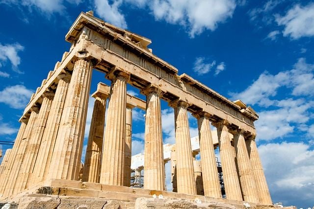 Veduta dal basso del Partenone sull’Acropoli di Atene, con le alte colonne doriche in pietra e parte della struttura superiore del tempio. Sullo sfondo, un cielo azzurro attraversato da nuvole bianche.