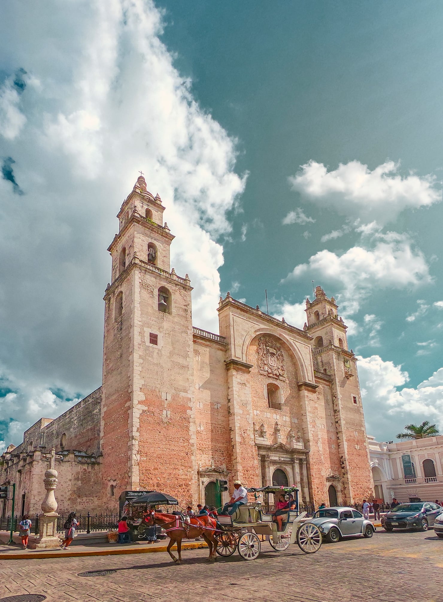 Low-angle view of a historic stone cathedral with two tall bell towers and an richly decorated façade. In front of the building, a horse-drawn carriage crosses the square, surrounded by people and cars.