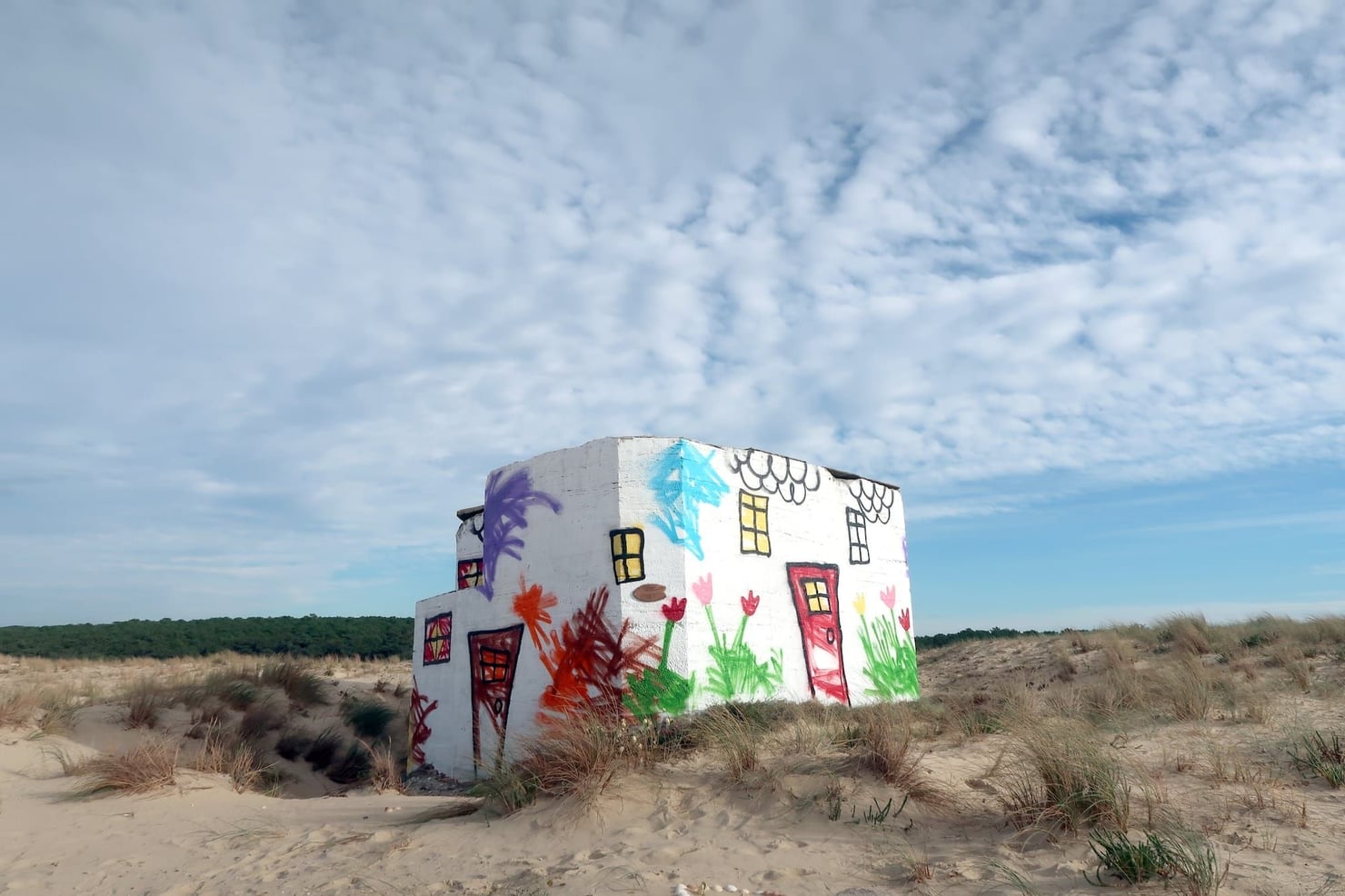 Piccola costruzione bianca tra le dune di sabbia, decorata con disegni colorati che raffigurano porte, finestre, fiori, piante e alberi. Intorno crescono ciuffi d’erba, sotto un ampio cielo azzurro attraversato da nuvole.