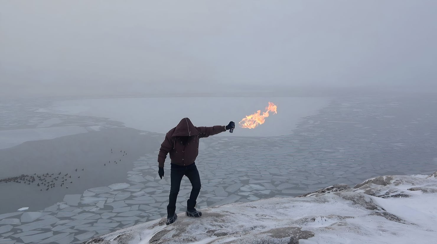 A hooded person stands on a snow-covered shore, extending one arm while holding a flame. In the background, a vast, partially frozen body of water stretches into the mist, with several birds visible on the left.