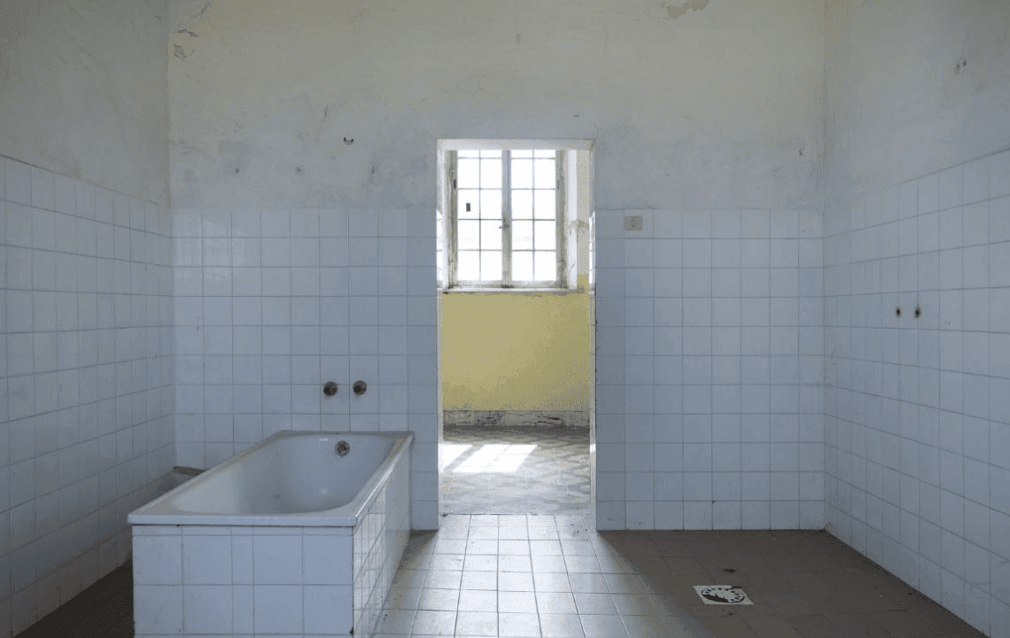 Interno di un ambiente spoglio e in disuso, con pareti rivestite di piastrelle bianche e una vasca rettangolare sulla sinistra. Al centro, una porta aperta conduce a una stanza illuminata da una grande finestra.