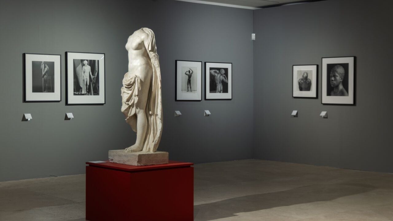 Sala espositiva con pareti grigie. Al centro, su un piedistallo rosso, è esposta una statua classica in marmo priva della testa e delle braccia, raffigurante una figura femminile drappeggiata. Sulle pareti circostanti sono disposte fotografie in bianco e nero di figure e corpi umani.