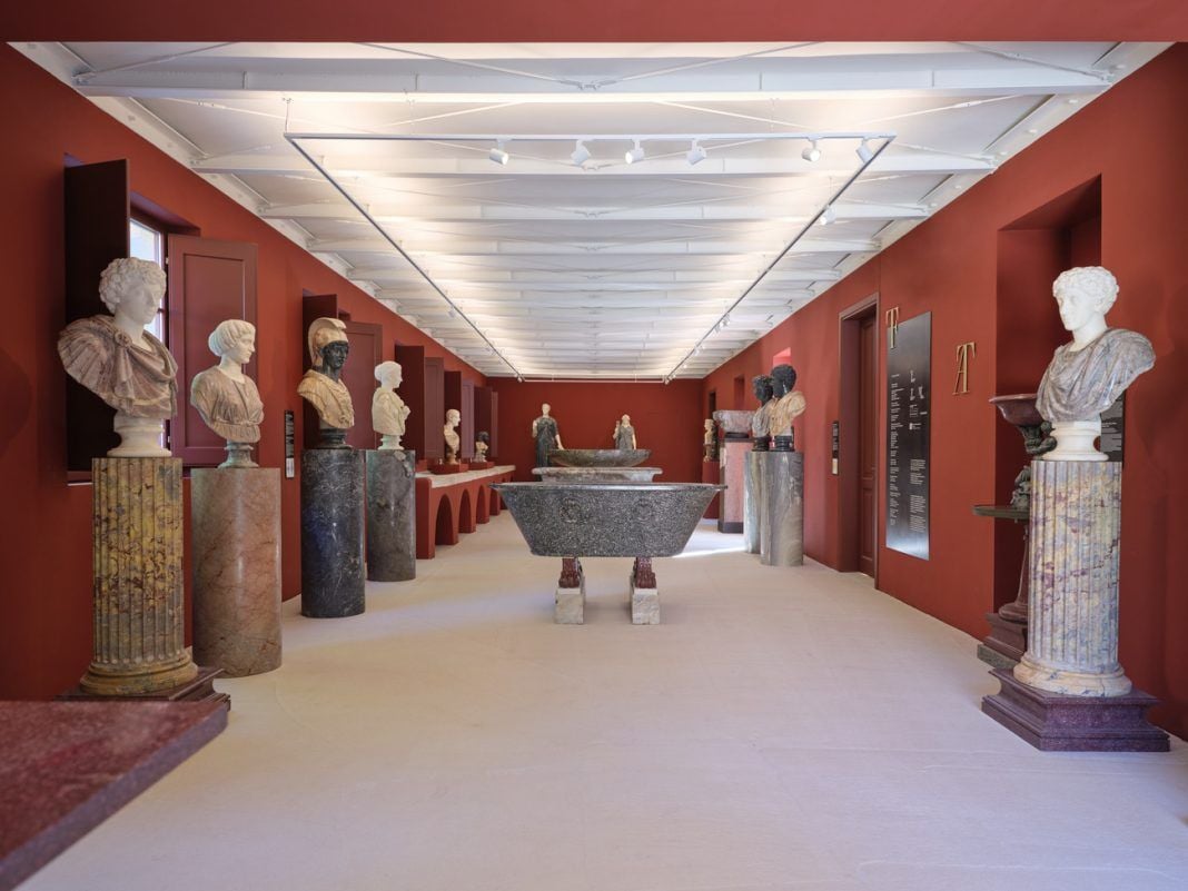 Interno dell’Antiquarium di Villa Albani Torlonia, con pareti rosse e una serie di sculture antiche esposte lungo il percorso. Busti e statue sono collocati su colonne e piedistalli in marmi diversi, mentre al centro della sala sono esposti grandi manufatti in pietra.