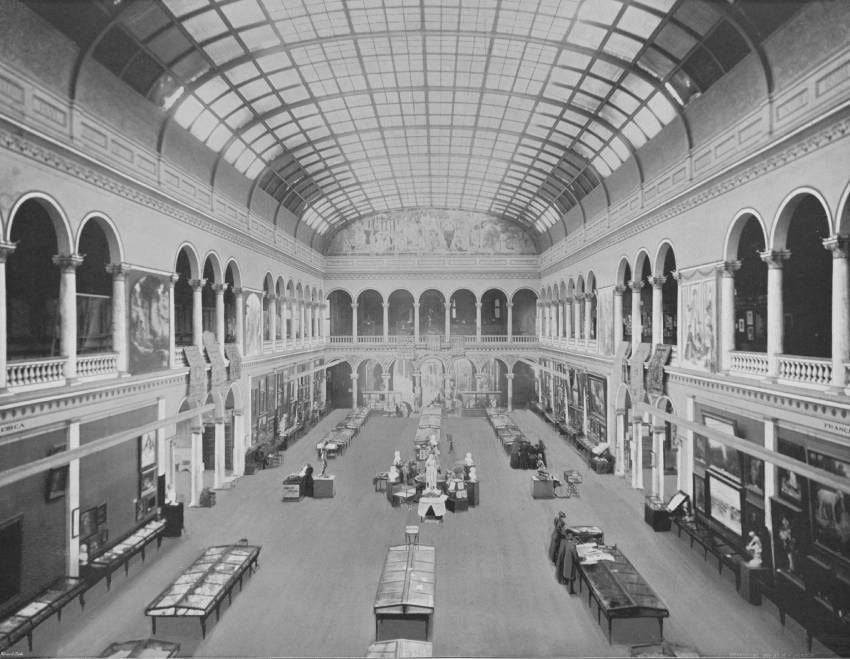 Fotografia storica in bianco e nero dell’interno di un grande museo, con un’ampia sala su due livelli scandita da arcate e coperta da una volta vetrata. Opere, sculture e teche espositive sono disposte lungo le pareti e al centro dello spazio.