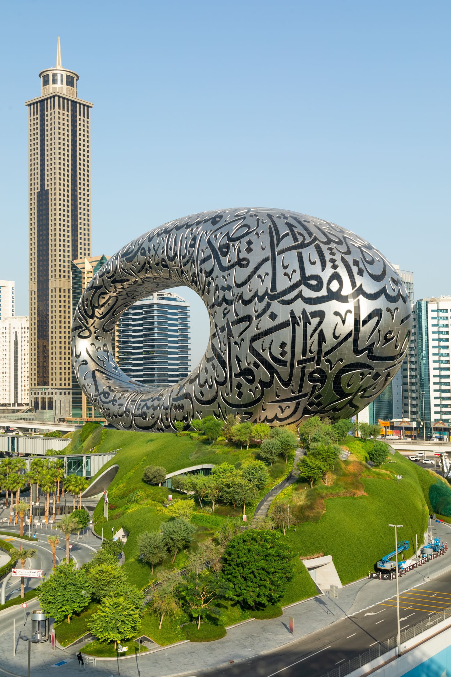 Veduta esterna del Museo del Futuro di Dubai, un grande edificio a forma di anello rivestito da pannelli metallici decorati con calligrafia araba. L’edificio sorge su una collina ricoperta di vegetazione, circondato da strade e grattacieli; sulla sinistra si innalza una torre alta e rettangolare.