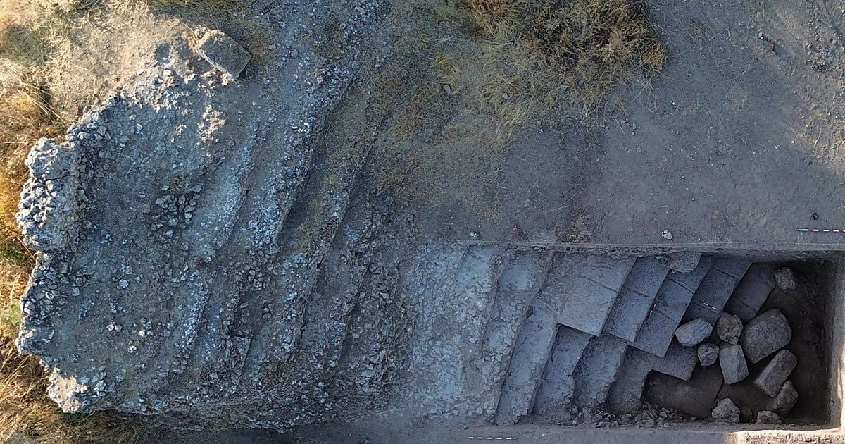 Veduta aerea di uno scavo archeologico, con resti di strutture murarie in pietra e tracce di antiche fondazioni visibili nel terreno. Sulla destra, un’area di scavo rettangolare mette in luce una struttura con gradini e blocchi di pietra.