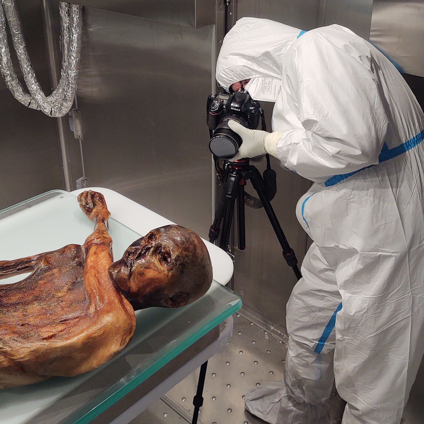 Un operatore in tuta protettiva fotografa una mummia umana all’interno di una struttura refrigerata, utilizzando una fotocamera montata su un treppiede.