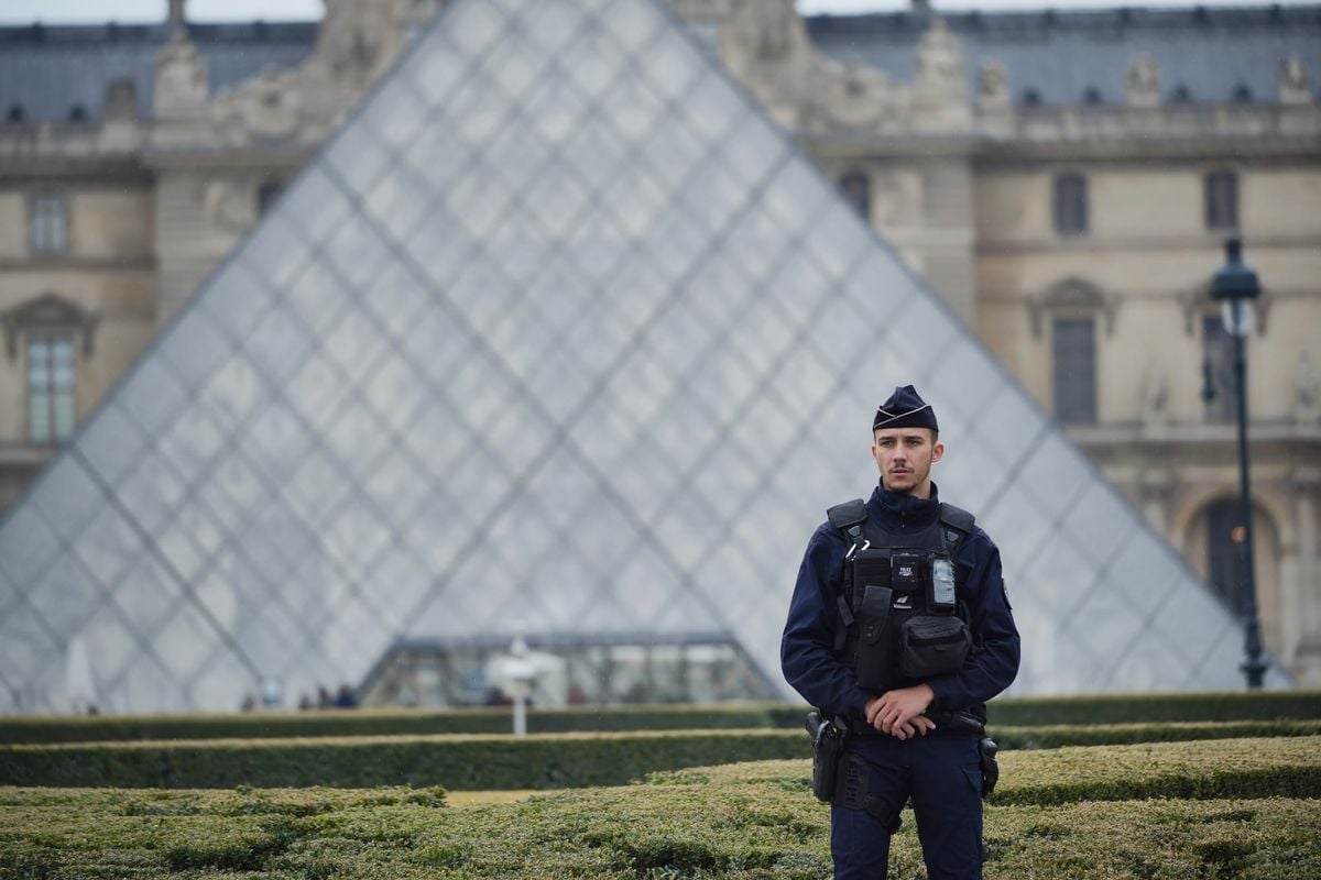 Un agente di sicurezza in uniforme è in piedi nei giardini del Louvre a Parigi. Sullo sfondo si vede la piramide di vetro del museo e, oltre i giardini, gli edifici storici del complesso.