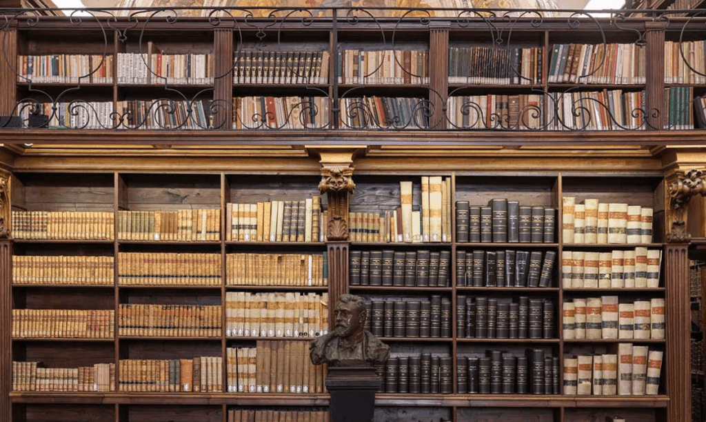 Interno di una biblioteca storica con scaffalature in legno riccamente decorate, colme di libri antichi e moderni. Al centro, davanti agli scaffali, è collocato un busto in bronzo su un piedistallo.