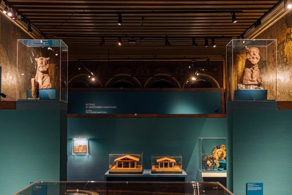 Allestimento della mostra “Etruschi e Veneti” a Palazzo Ducale di Venezia, con reperti archeologici esposti in teche e su piedistalli. Al centro dell’allestimento sono visibili due modellini di edifici antichi, mentre ai lati sono esposte due sculture in pietra.