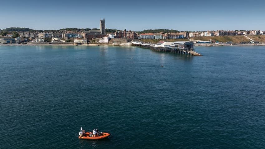 Veduta panoramica di una città costiera affacciata sul mare, con edifici e una grande chiesa lungo il litorale e un molo che si protende sull’acqua. In primo piano, una piccola imbarcazione arancione con alcune persone a bordo.
