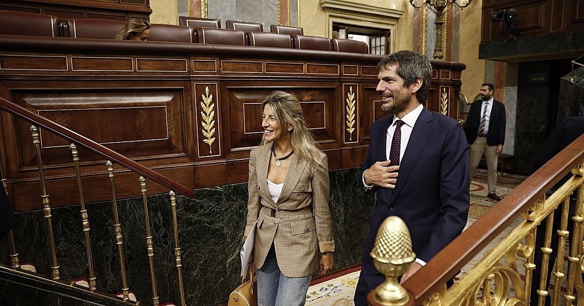 Una donna e un uomo in abiti formali camminano all’interno di un’aula parlamentare, accanto ai banchi in legno decorati con elementi dorati. Entrambi sorridono; sullo sfondo sono visibili altre persone e i sedili dell’aula.