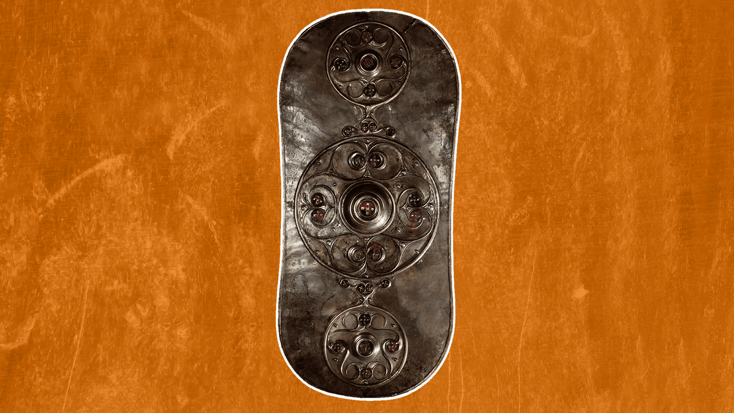 Oggetto metallico antico di forma allungata e arrotondata, decorato con tre motivi circolari disposti verticalmente. Le decorazioni presentano elementi geometrici e piccoli inserti rossastri. Il reperto è mostrato su uno sfondo arancione.