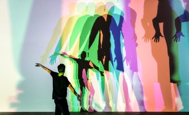 Una persona, con la propria ombra ripetuta in diversi colori, si trova davanti a una parete sulla quale sono proiettate altre ombre multicolori.