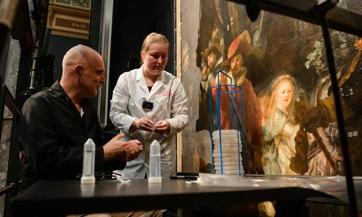 Due persone lavorano al tavolo di un laboratorio di restauro davanti a un grande dipinto. Una indossa un camice bianco e tiene in mano uno strumento, mentre l’altra, vestita di nero, osserva il lavoro. Sul tavolo sono presenti strumenti e materiali per il restauro; sullo sfondo si vede il dipinto.