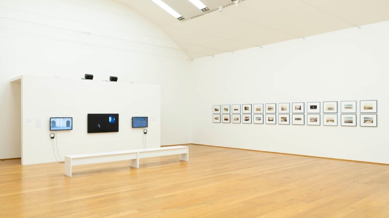 Sala espositiva di un museo con pareti bianche e pavimento in legno. Sulla parete a sinistra sono installati tre schermi, mentre sulla parete di fondo è esposta una serie di fotografie o immagini incorniciate disposte in due file. Due lunghe panche bianche sono collocate davanti agli schermi.