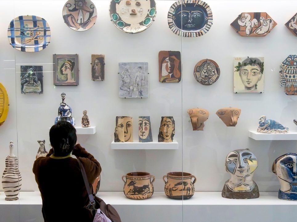 Vetrina museale con una collezione di opere in ceramica decorate, tra piatti, pannelli, vasi e sculture. Le opere presentano motivi geometrici e figure umane e animali in diversi colori. In primo piano, una persona osserva la collezione.