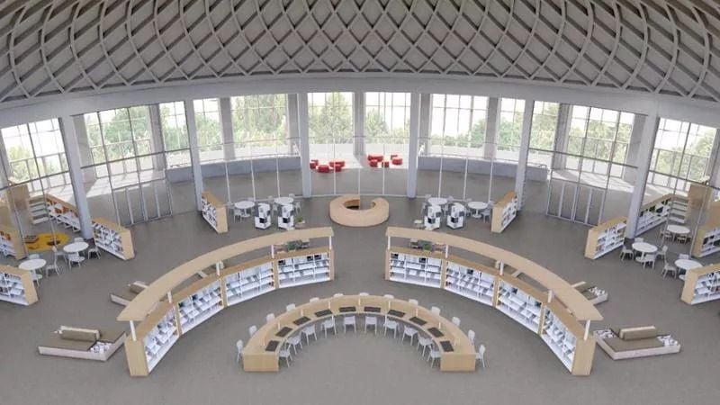 Veduta dall’alto dell’interno di una biblioteca moderna, organizzata in uno spazio ampio e luminoso con grandi finestre e soffitto a volta. Scaffali curvi disposti a semicerchio circondano un’area centrale con tavoli, sedie e postazioni di lavoro.