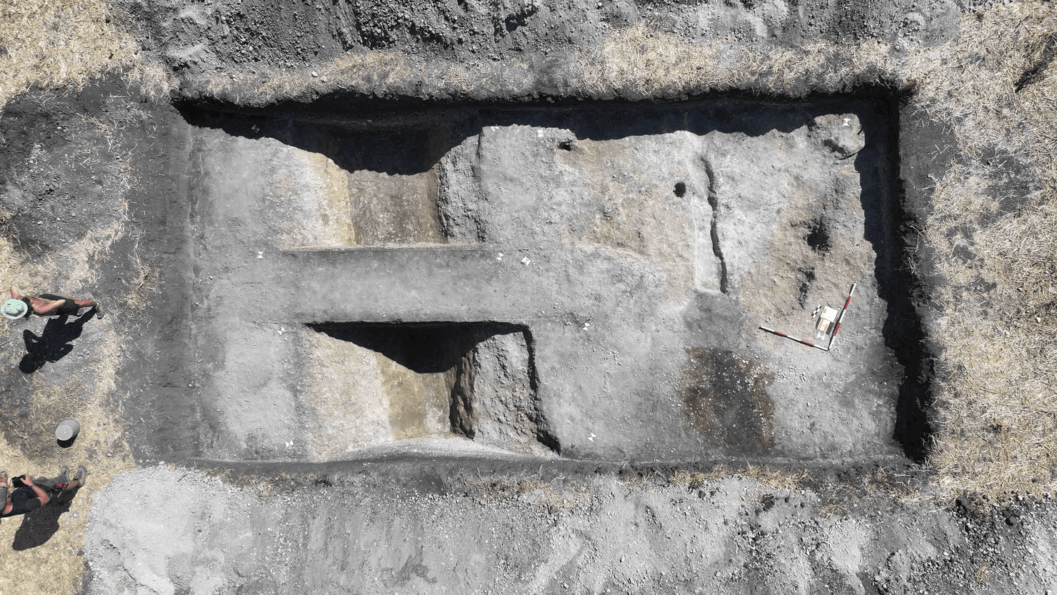 Veduta aerea dall'alto di uno scavo archeologico di Chleby di forma rettangolare nel terreno, con fossati interni e due archeologi visibili sul lato sinistro.